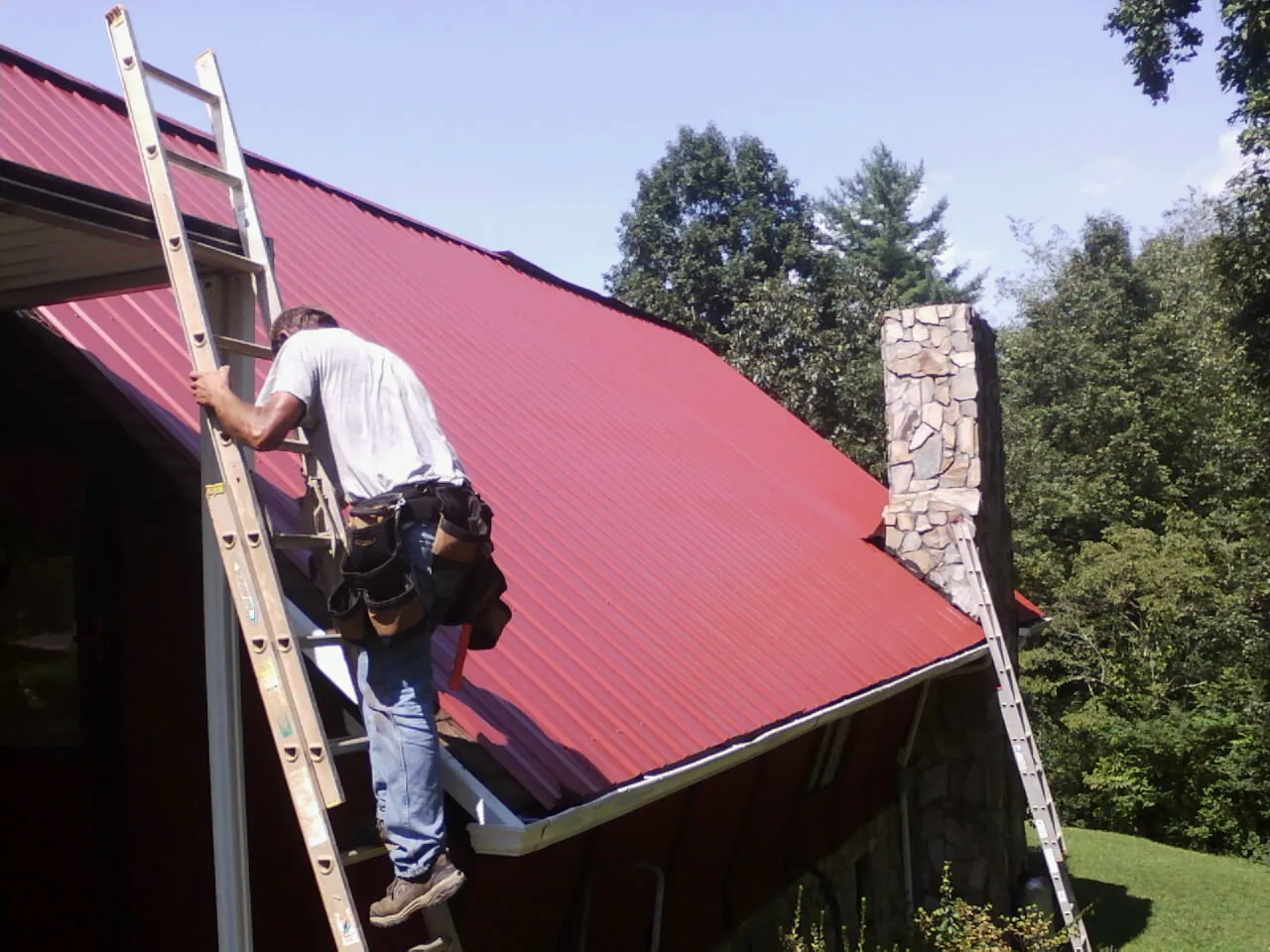 Roofer on a ladder beside a red metal roof and stone chimney — Soffit & Fascia Repair by Roofing Brothers in Darien